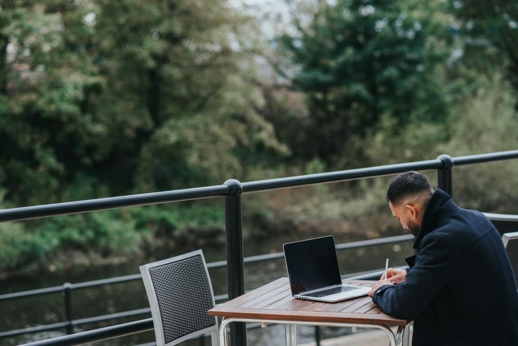 anonymous ethnic worker writing near laptop on cafe terrace